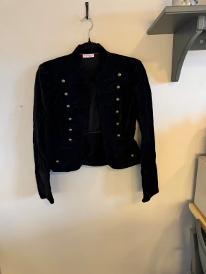 Svart sammetskavaj från Atmosphere - Stilren svart sammetskavaj från Atmosphere med militärinspirerad design och dubbla rader av dekorativa guldknappar framtill. Kortare modell med markerade axlar och klassisk krage. Perfekt för dig som vill sticka ut med en cool och elegant look.