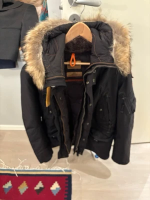 Svart parkas från Parajumpers - Säljer en svart parkas från Parajumpers Masterpiece Series med avtagbar fuskpäls på huvan, flera fickor och orange detalj vid halsen. Jackan har en klassisk parkaslook och är perfekt för kalla dagar. Slim fit-modell med snygga sömdetaljer.