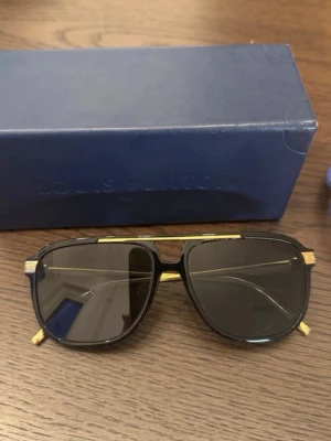 Louis Vuitton svarta solglasögon guld - Snygga solglasögon från Louis Vuitton med svart båge och detaljer i guld. Glasen är mörka och modellen har en klassisk, fyrkantig form med exklusiv känsla. Perfekt accessoar för att lyfta din stil.