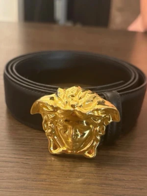 Svart skärp med guldspänne från Versace - Snyggt svart skärp från Versace med ikoniskt guldigt Medusa-spänne. Bältet är gjort i slätt skinn och har en klassisk, stilren design som lyfter vilken outfit som helst. Perfekt accessoar för dig som gillar exklusiva detaljer.