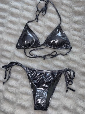 Glansig silvrig bikini med knyt - Snygg silvrig bikini i glansigt material med trekantsöverdel och knyt både i ryggen och vid höfterna. Perfekt för dig som vill sticka ut på stranden eller vid poolen. Klassisk modell med trendig finish. Köpt på amazon och aldrig använd. Storlek S 