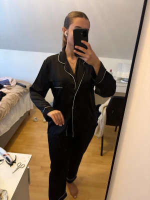 Svart satinpyjamas med vita detaljer - Snygg svart pyjamas i satin med vita kontrastsömmar. Setet består av långärmad skjorta med krage och knäppning framtill samt matchande byxor. Perfekt för mysiga kvällar hemma. Se defekt sista bild översta och nedersta knappen saknas. 