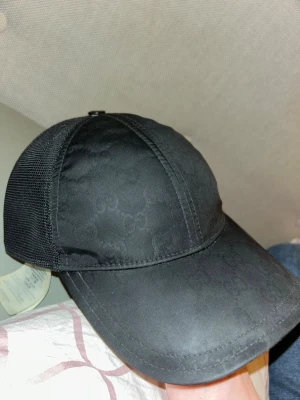 Svart keps från Gucci med meshdetaljer - Snygg svart keps från Gucci med diskret GG-mönster på framsidan och meshpaneler på sidorna och baksidan för extra ventilation. Justerbar snapback-stängning bak och klassisk böjd skärm. Perfekt för dig som vill ha en stilren och exklusiv accessoar.