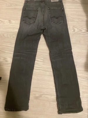 Grå jeans från Replay - Säljer ett par grå jeans från Replay med klassisk femficksdesign. Jeansen har snygga slitningar och kontrastsömmar, samt Replay-logga på bakfickan. Perfekta för en avslappnad och trendig look.