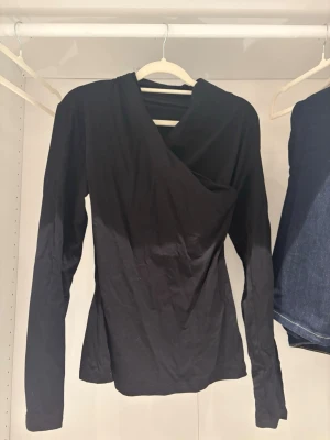 Svart omlottopp med lång ärm - Stilren svart omlottopp med lång ärm och asymmetrisk v-ringning. Toppen har en figurnära passform och är tillverkad i mjukt bomullsmaterial. Perfekt för dig som gillar minimalistisk och trendig stil.