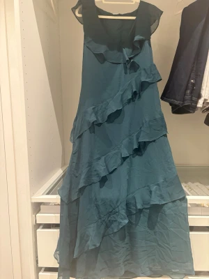 Grön långklänning med volanger - Snygg grön långklänning med flera lager av volanger och ärmlös design. Klänningen har en luftig och flowy känsla, perfekt för varma dagar. Den har en enkel men elegant look med V-halsringning.