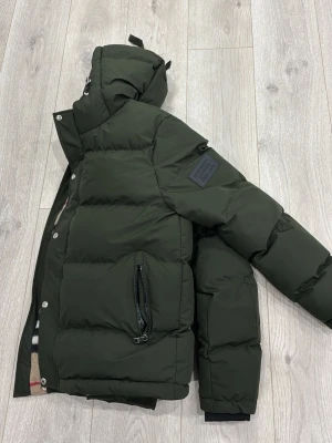 Mörkgrön pufferjacka Burberry - Snygg mörkgrön pufferjacka från Burberry med stor huva och patch med logga på ärmen. Jackan har dragkedja och tryckknappar framtill samt en praktisk ficka med dragkedja. Insidan har klassiskt Burberry-mönster. Perfekt för kalla vinterdagar. Den har använts 5-6 gånger annars har den legat i garderoben, vill ändra min stil och denna passar inte den stilen så jac vill gärna få den sålt om, ni är intresserade hör av er gärna