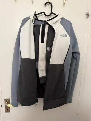 The North Face hoodie i grått och vitt - Snygg hoodie från The North Face i färgblockad design med ljusgrå, mörkgrå och vit. Dragkedja framtill, huva och logga på bröstet. Tillverkad i mjukt material som känns bekvämt och passar perfekt till en avslappnad stil.