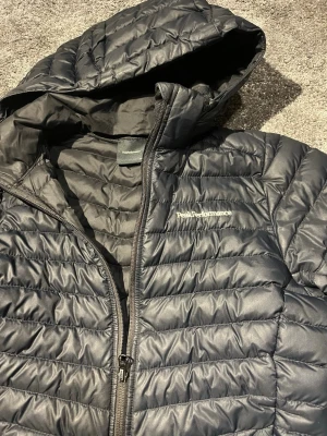 Svart dunjacka Peak Performance - Snygg svart dunjacka från Peak Performance med quiltad design och dragkedja framtill. Jackan har en praktisk huva och diskret logga på bröstet. Perfekt för kalla dagar och enkel att matcha med olika outfits.