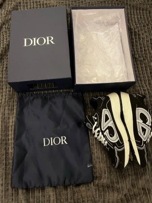 Svarta Dior B22  - Säljer ett par svarta Dior B22 sneakers med vita och grå detaljer. Skorna har chunky sula, vita skosnören och coola kontrastmönster på sidan. Kommer med originalkartong och dustbag. Perfekta för dig som gillar exklusiva och trendiga sneakers.