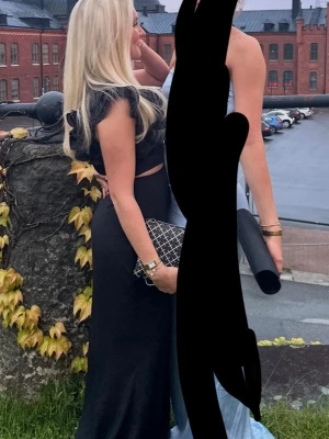 Balklänning  - Snygg svart långklänning med volangdetalj på axeln och liten cut-out vid midjan. Klänningen har en elegant och figurnära passform, perfekt för speciella tillfällen.