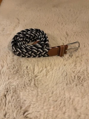 Svartvitt flätat bälte med läderdetaljer - Snyggt flätat bälte i svart, vitt och grått med bruna läderdetaljer vid spännet. Bältet har ett silverfärgat metallspänne och passar perfekt till jeans eller chinos för en stilren look.