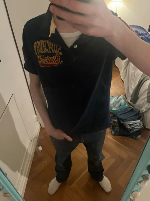 Mörkblå pikétröja från Superdry - Säljer en mörkblå pikétröja från Superdry med broderad gul och röd logga på bröstet samt siffran 68 i gult och rött på ärmen. Klassisk krage med knappar och korta ärmar. Perfekt för en avslappnad och sportig stil. Obs den ät sydd längst nere på tröjan (det är bra sytt och borde ej gå up i tvätten)