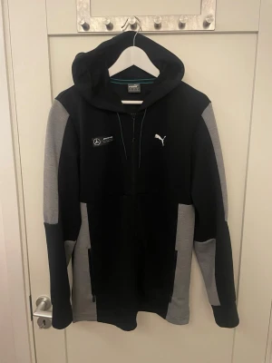 Svart och grå Puma Mercedes hoodie - Snygg svart hoodie från Puma med grå ärmar och detaljer. Framtill finns Mercedes AMG Petronas Motorsport-logga och Puma-logga. Dragkedja hela vägen, huva med snören och två fickor. Perfekt för dig som gillar motorsport och streetwear.