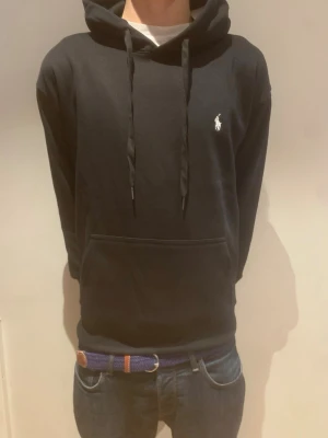 Svart hoodie från Ralph Lauren - Pris går att diskuteras!! Snygg svart hoodie från Ralph Lauren med klassisk huva och dragsnören. Har en vit broderad logga på bröstet och en stor magficka framtill. Tillverkad i mjukt material som känns skönt mot huden. Perfekt för en avslappnad och stilren look. 