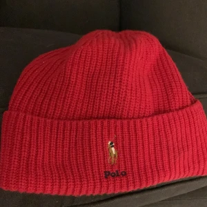 Röd ribbstickad mössa från Ralph lauren - Snygg röd ribbstickad mössa från Ralph Lauren med uppvikt kant och den klassiska broderade polospelaren framtill. Perfekt accessoar för att hålla värmen och addera lite färg till din outfit.