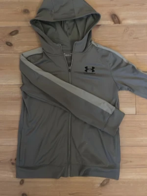 Grå hoodie från Under Armour - Snygg grå hoodie från Under Armour med dragkedja framtill och huva. Tillverkad i ett mjukt och sportigt material, perfekt för en aktiv stil. Loggan är tryckt i svart på bröstet och ärmarna har en ljusare grå detalj längs sidan.