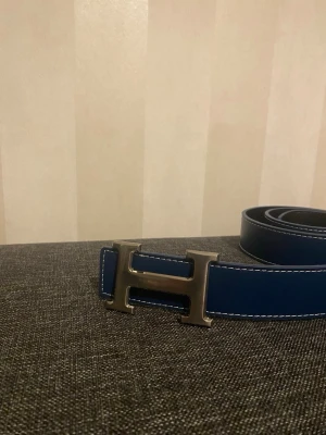 Blått skärp med H-spänne - Snyggt blått skärp med ett stort, silverfärgat H-spänne. Skärpet har vita sömmar längs kanterna och en stilren design som sticker ut. Perfekt accessoar för att lyfta din outfit.