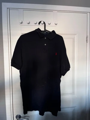 Svart pikétröja från Polo Ralph Lauren - Klassisk svart pikétröja från Polo Ralph Lauren med korta ärmar och krage. Tröjan har knappar framtill och den ikoniska röda logotypen broderad på bröstet. Tillverkad i mjuk bomull som är skön att bära.
