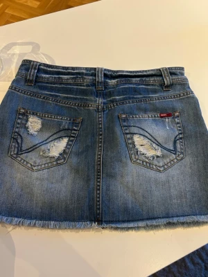 Blå jeanskjol med slitningar från Guess - Trendig blå jeanskjol från Guess med slitna detaljer och fransad nederkant. Kjolen har klassiska fickor fram och bak samt hällor för bälte. Perfekt för dig som gillar en avslappnad och cool stil.