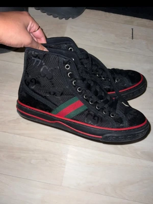 Svarta Gucci sneakers med röd/grön rand - Snygga svarta high top sneakers från Gucci med klassiska GG-mönstret över hela skon och ikoniska röd-gröna ränder på sidan. Skorna har svarta snören, metallöljetter och en svart sula med röd detalj runt om. Perfekta för dig som vill sticka ut.