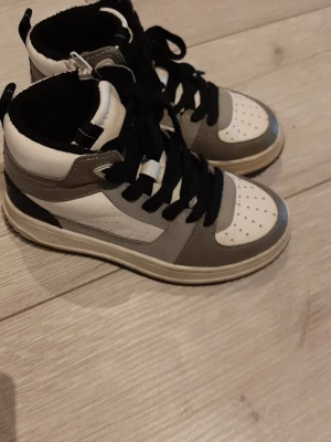 Gråvita high top sneakers för barn  - Snygga high top sneakers för barn  i grått och vitt med svarta skosnören. Skorna har perforerad tå och klassisk sportig design. Perfekta för dig som gillar streetstyle och vill ha en cool look.