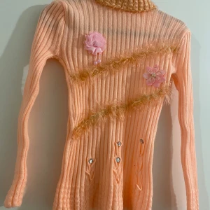 Ljus orange ribbad polotröja med blommor - Säljer en ljus orange ribbad polotröja med lång ärm och hög krage. Tröjan har dekorativa blommor i rosa och pärldetaljer framtill samt fluffiga guldfärgade ränder. Materialet är stickat och ger en mysig känsla. Perfekt för dig som gillar unika detaljer och färg.