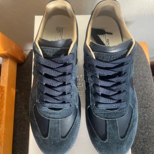 Maison Margiela blå sneakers mocka/läder - Snygga sneakers från Maison Margiela fast i mörkblå mocka och läder! Klassisk siluett med rund tå, platt sula och matchande blå snörning. Insidan är beige för en clean kontrast. Perfekta för dig som gillar stilrena skor. Box kommer med.