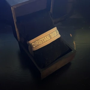 Grått Christian Dior armband - Snyggt grått armband från Christian Dior med texten 'J'Adior' broderad i vitt. Armbandet har justerbar knytning med tofsar i ändarna och en stilren, vävd design som känns både lyxig och trendig. Perfekt accessoar för att lyfta din outfit.