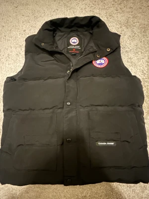 Svart dunväst från Canada Goose M - Svart dunväst från Canada Goose med klassisk Arctic Program-patch på bröstet. Västen har hög krage, två stora fickor framtill och knäppning med både dragkedja och tryckknappar. Perfekt för kalla dagar och har en clean, stilren look.