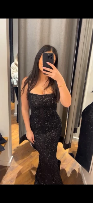 Svart glittrig långklänning - Säljer en svart långklänning med glittriga paljetter och figurnära passform. Klänningen är axelbandslös med rak urringning och har en elegant siluett som framhäver midjan. Perfekt för bal eller fest. Materialet känns lyxigt och skimrar snyggt i ljuset.