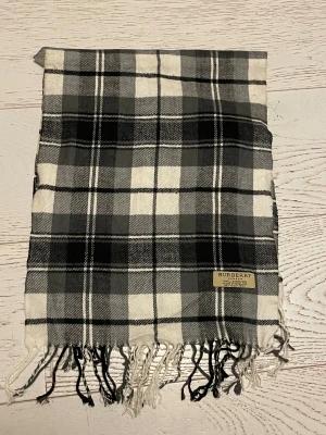 Rutig halsduk från Burberry i ull - Snygg halsduk från Burberry i klassiskt rutigt mönster med svarta, gråa och vita toner. Gjord av 100% lammull och har fransar i ändarna. Perfekt längd för att vira flera varv och mjuk känsla mot huden. Märkt med Burberry London-tag.