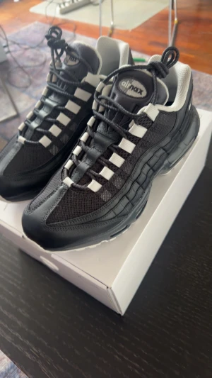 Nike Air Max 95 svart/vit sneakers - Nike Air Max 95 sneakers i svart och vitt med ikonisk vågig design och synlig Air-dämpning i sulan. Ovandel i mesh och skinn med vita detaljer och snörning. Perfekta för dig som vill ha en klassisk och sportig look med maximal komfort.