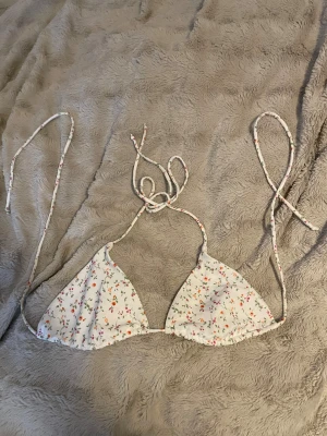 Vit blommig bikiniöverdel med snörning - Supersöt bikiniöverdel i vit med små blommor i rosa, orange och grönt. Klassisk trekantsmodell med tunna snören att knyta i nacken och ryggen. Perfekt för strandhäng och soliga dagar. Materialet känns mjukt och följsamt mot huden.