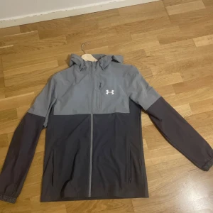 Grå vindjacka från Under Armour - Snygg tvåfärgad vindjacka från Under Armour i ljusgrått och mörkgrått. Jackan har huva, hel dragkedja framtill och en diskret logga på bröstet. Långärmad modell med praktiska fickor och sportig look, perfekt för utomhusaktiviteter.