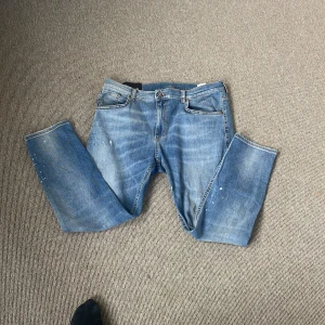 Dondup jeans - Säljer ett par klassiska blå jeans från Dondup med raka ben och femficksmodell. Jeansen har en ljus tvätt och diskreta slitningar samt små vita färgstänk för en cool, avslappnad look. Passar perfekt till vardagsstilen.
