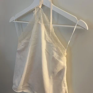 Champagnefärgad spetsblus i satin - Säljer en champagnefärgad blus med tunna axelband och vacker spetsdetalj längs halsringningen. Blusen är gjord i glansigt satinmaterial och har en elegant, ärmlös design som ger en lyxig känsla.