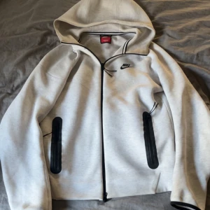 Nike tech fleece kofta - ”nypris 1600” Nike hoodiejacka i ljusgrått med svart dragkedja och svarta detaljer vid fickorna. Jackan har huva, lång ärm och liten Nike-logga på bröstet. Tillverkad i mjuk bomullsblandning, perfekt för chill eller träning.