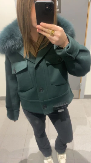 Mörkgrön jacka med päls från meotine  - Snygg mörkgrön jacka med stor fluffig päls runt kragen. Jackan har en boxig form, stora fickor med lock och knappar framtill. Materialet är tjockt och känns lyxigt, perfekt för kalla dagar. Pälsen ger en cool och trendig vibe.
