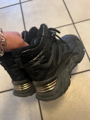 Svarta chunky boots med pälsfoder - Chunky svarta boots med grov sula och metallic-detalj på hälen. Skorna har snörning och är fodrade med mjuk svart päls på insidan för extra värme. Yttermaterialet är syntet med glansig finish och robust design, perfekt för dig som gillar streetstyle.