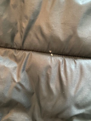 Svart pufferjacka Super Dry - Svart pufferjacka från Superdry MTN Sports Puffer Series med broderad logga på bröstet. Jackan har huva, två fickor med dragkedja framtill. Perfekt för kalla dagar. Det finns små hål på jackans rygg. Se bild. 