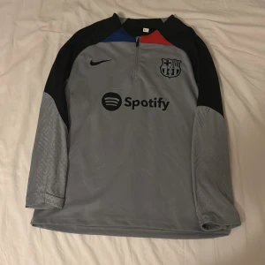 Grå långärmad FC Barcelona tröja - Säljer en grå långärmad FC Barcelona fotbollströja från Nike med svart logga och Spotify-tryck på bröstet. Tröjan har svarta axelpartier, blå och röd detalj vid halsen samt subtilt mönster på ärmarna. Perfekt för dig som vill representera Barca.
