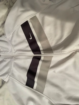 Vit vindjacka från Nike med ränder - Säljer en fräsch vit vindjacka från Nike med coola svarta och gråa ränder över bröstet. Jackan har hel dragkedja, ribbad krage och muddar samt två sidofickor. Perfekt för dig som gillar sportig stil och vill ha något som sticker ut.
