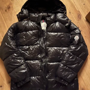 Svart Moncler pufferjacka med huva - Svart pufferjacka från Moncler med glansig finish, huva och klassisk logga på ärmen. Jackan har dragkedja framtill och quiltad design som ger en schysst volym. Perfekt för kalla dagar och har coola detaljer som serietikett på insidan.