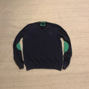 Mörkblå v-ringad tröja Riley S - Snygg mörkblå v-ringad tröja från Riley i storlek S. Tröjan har gröna detaljer på armbågarna och insidan av kragen samt ett broderat grönt emblem på bröstet. Perfekt för dig som gillar stilrena och unika detaljer.