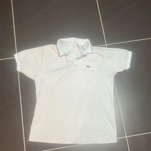 Ljusblå pikétröja från Lacoste M - Klassisk  pikétröja från Lacoste i storlek M med broderad krokodil på bröstet. Tröjan har korta ärmar, krage och knappar framtill. 