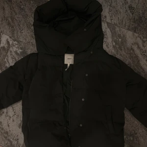 Mörkgrön pufferjacka från Object - Svart pufferjacka från Object med stor, mysig huva och dragkedja framtill. Jackan har knappar och två fickor på sidorna. Perfekt för kalla dagar och har en schysst, lite oversized passform. En knapp är sönder men jackan har en dragkedja också så inget som syns 