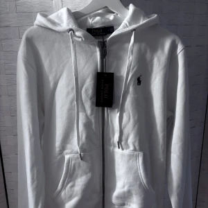 Vit hoodie från Polo Ralph Lauren, oanvänd  - Vit hoodie från Polo Ralph Lauren med dragkedja framtill, snörning i huvan och klassisk logga broderad på bröstet. Tröjan har två fickor och är tillverkad i mjuk bomull. Oanvänd då den inte passade och jag fick den som present!! Ord pris: strax över 2000kr
