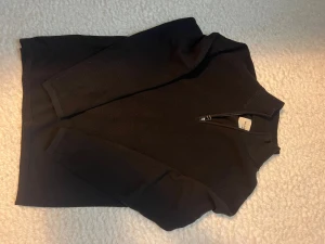 Svart stickad half zip-tröja från Marcus  - Svart stickad tröja från Marcus med half zip-dragkedja vid halsen. Tröjan har långärmad design och ribbade muddar vid ärmslut och hals. Perfekt för dig som gillar stilrena och klassiska plagg med en modern touch.