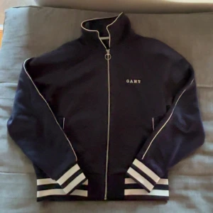 Svart track jacket från GANT, strl L - Svart track jacket från GANT med vita detaljer och dragkedja framtill. Jackan har höga muddar med svarta och vita ränder, ståkrage och broderad GANT-logga på bröstet. Perfekt för en sportig och clean look.